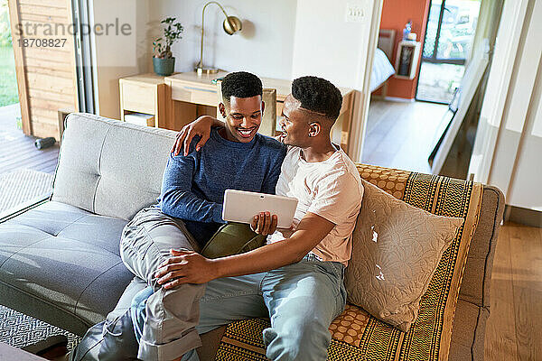 Glückliches junges schwules männliches Paar  das sich auf dem Sofa umarmt und ein digitales Tablet benutzt