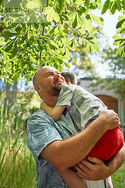 Glücklicher Vater hält und umarmt seinen Sohn unter einem Baum im sommerlichen Hinterhof