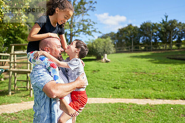 Vater hält süße  verspielte Kinder im sonnigen Sommerpark