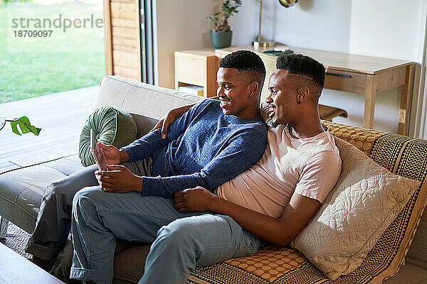 Glückliches junges schwules männliches Paar mit digitalem Tablet auf dem Sofa im Wohnzimmer