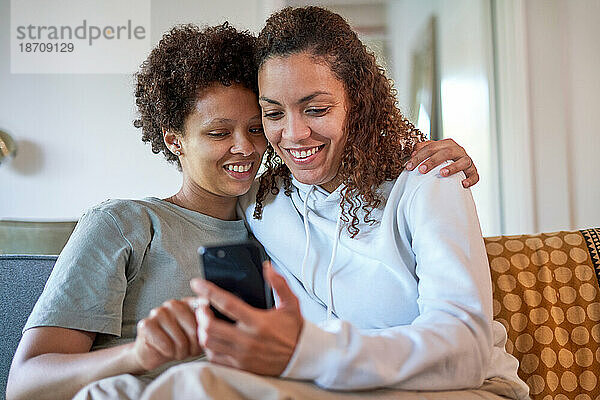 Glückliches lesbisches Paar umarmt sich und nutzt sein Smartphone auf dem heimischen Sofa