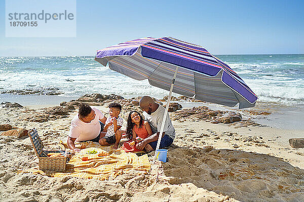 Schwules männliches Paar und Kinder essen unter Sonnenschirm am sonnigen Strand zu Mittag