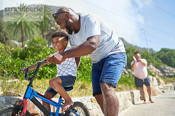Vater bringt seinem glücklichen Sohn bei  wie man auf der sonnigen Promenade Fahrrad fährt
