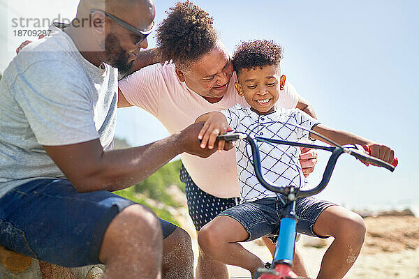 Glückliches schwules männliches Paar hilft seinem Sohn beim Fahrradfahren am sonnigen Strand