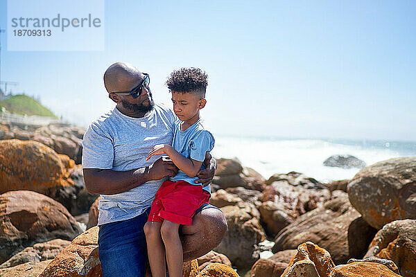 Zärtlicher Vater hält Sohn auf sonnigen Strandfelsen