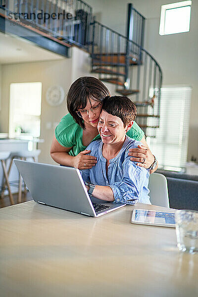 Glückliches lesbisches Paar  das gemeinsam am Esstisch zu Hause einen Laptop benutzt