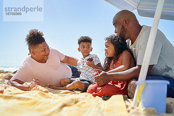 Schwules männliches Paar und Kinder essen auf einer Decke am sonnigen Strand