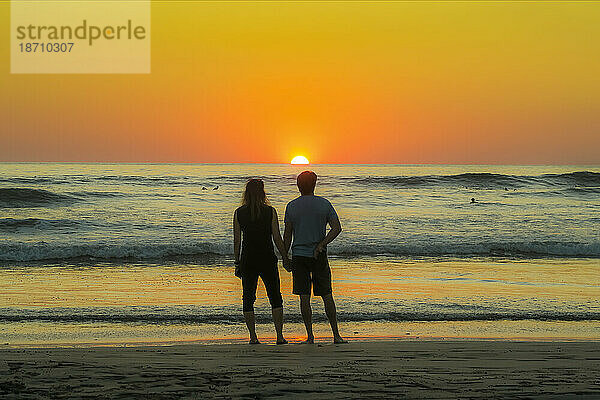 Ein Paar hält Händchen am Strand von Guiones  wo sich Menschen zum Surfen und Beobachten des Sonnenuntergangs versammeln  Playa Guiones  Nosara  Guanacaste  Costa Rica  Mittelamerika