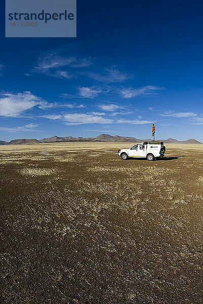 Mann steht auf dem Dach eines Expeditionslastwagens mitten in der afrikanischen Wüste. Weite Weite und blauer Himmel.