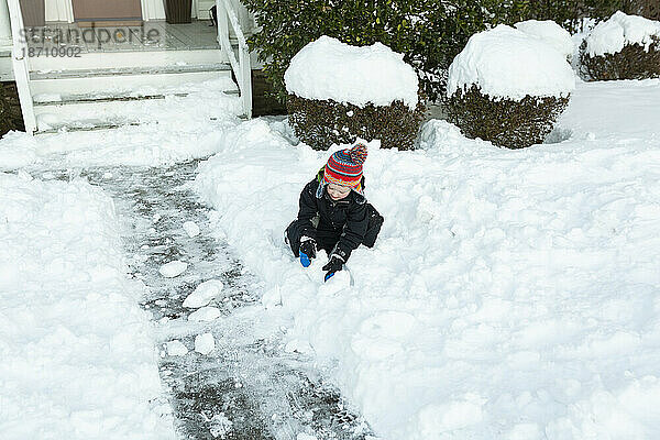 Junge im Vorschulalter mit buntem Hut spielt im Schnee vor dem Haus