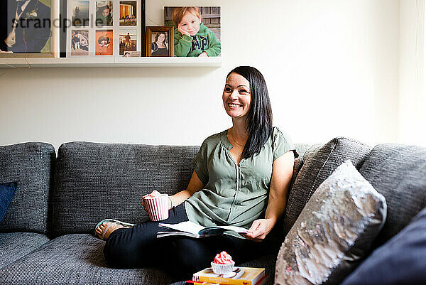 Eine Frau lächelt zu Hause auf dem Sofa  während sie liest und sich etwas Gutes tut