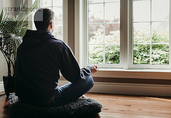 Mann sitzt drinnen auf dem Boden und meditiert vor Fenstern.