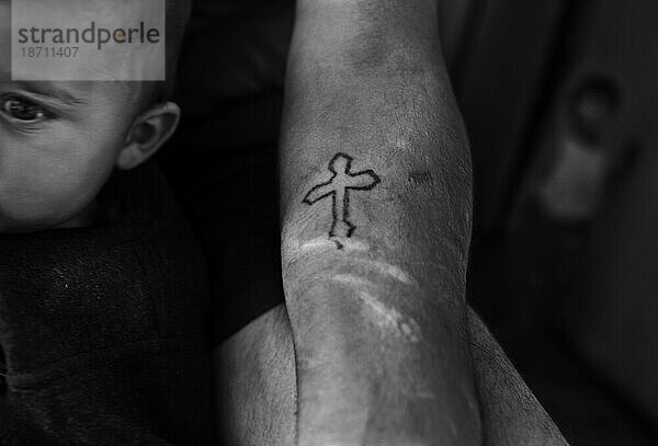 Ein Kruzifix-Tattoo auf dem Arm eines Mannes  der ein kleines Kind hält.