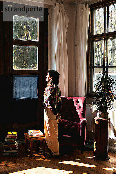 Frau schaut zu Hause aus dem Fenster  mit Sonne im Gesicht und Büchern