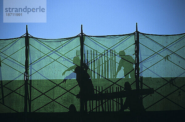 Silhouetten und Schatten von zwei Bauarbeitern hinter einer grünen Maschenbarriere auf einer Baustelle in Singapur.