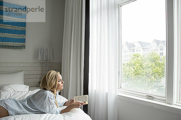 Blondine liest ein Buch und blickt aus dem Fenster