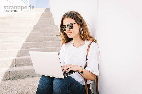 Vorderansicht einer glücklichen Studentin  die mit einem Laptop auf der Treppe einer Universität arbeitet