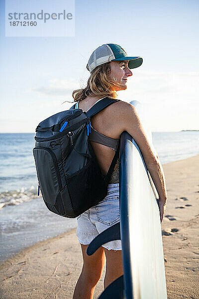 Frau spaziert bei Sonnenuntergang mit Surfbrett und Rucksack am Strand entlang