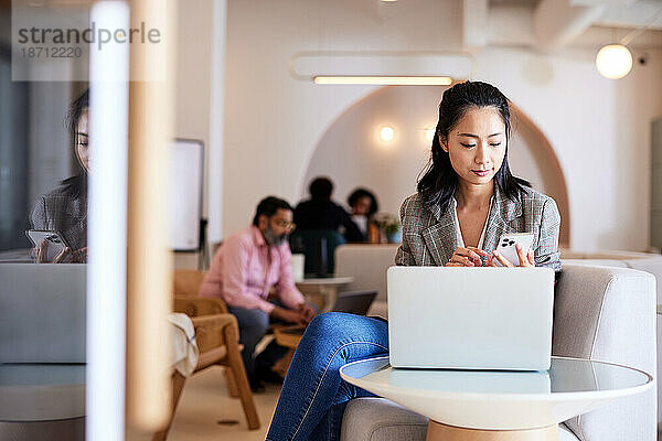 Geschäftsfrau sitzt mit Laptop am Tisch im Café und benutzt Smartphone
