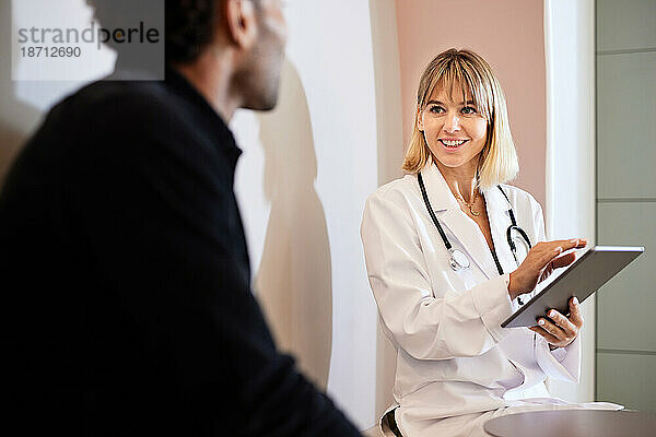 Blonde Ärztin hält Tablette in der Hand  während sie mit einem männlichen Patienten spricht