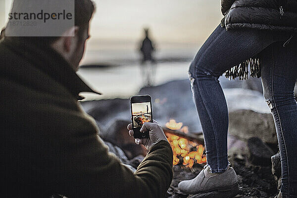 Ein Mann fotografiert mit seinem Handy oder Smartphone ein Lagerfeuer