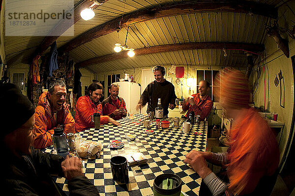 Sieben Männer in Skikleidung hängen nachts in einer rustikalen Hütte herum  trinken  spielen Karten und lachen.
