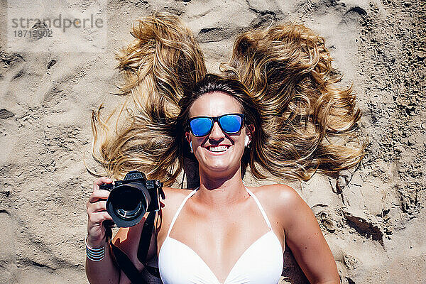Nahaufnahme einer blonden Frau mit Sonnenbrille  die im Strandsand liegt