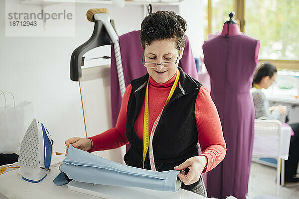 Ältere und fröhliche Frau bügelt in einem Modedesignstudio