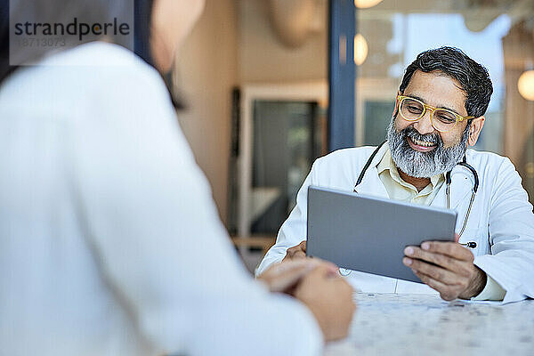 Glücklicher männlicher Arzt  der einen Tablet-PC verwendet und mit einer Patientin sitzt