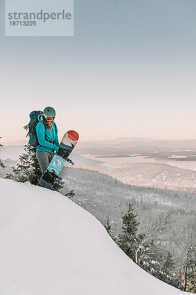 Frau mit Skibrille und Snowboard auf einem Berg mit Blick auf den See