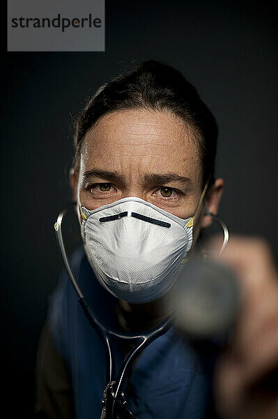 Studioporträt einer 42-jährigen kaukasischen Krankenschwester  die eine N95-Maske trägt und ein Stethoskop auf die Kamera richtet.