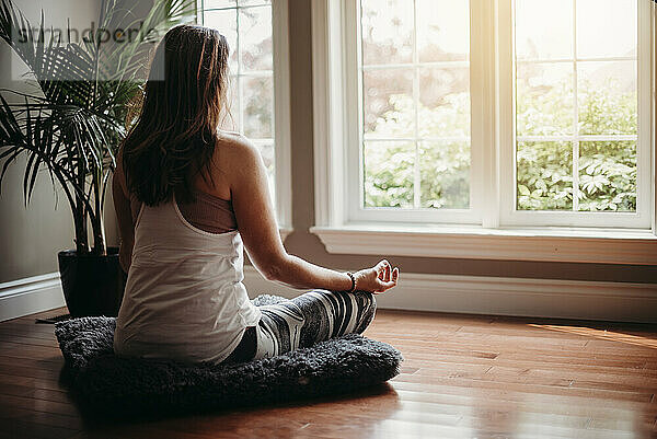 Frau sitzt drinnen auf dem Boden und meditiert vor Fenstern.