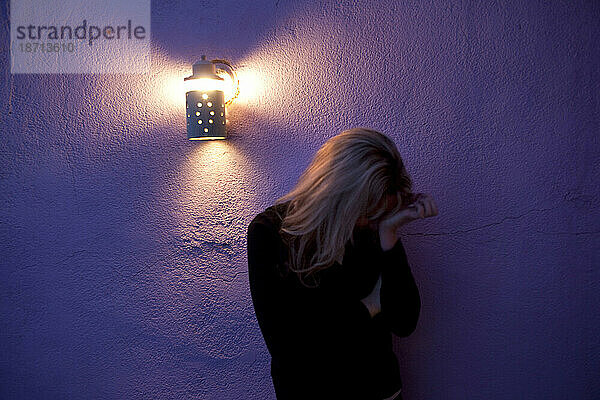 Eine Frau steht nachts mit dem Kopf in den Händen vor einer violetten Wand.