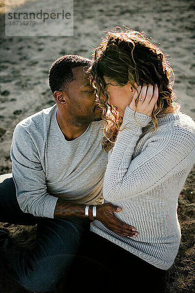 Mann und Frau umarmen sich  während ihr Mann am Strand den Bauch der schwangeren Frau berührt