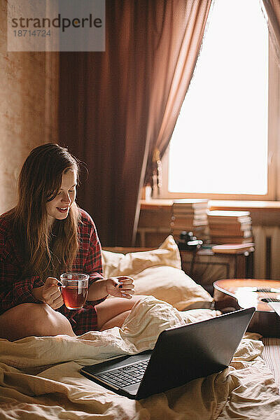 Junge Frau trinkt Tee im Bett und sitzt vor dem Laptop