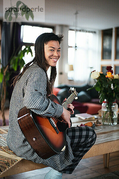 Hübsche asiatische Frau lächelt  während sie in einem schönen Zuhause Gitarre spielt