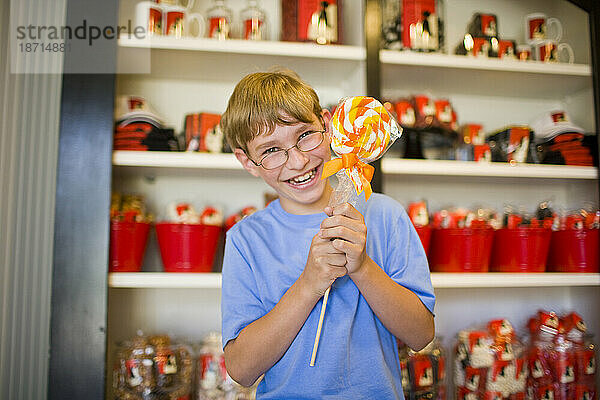 Ein glücklicher kleiner Junge mit Brille lächelt in die Kamera und hält einen riesigen Lutscher in einem Süßwarenladen in Kalifornien.