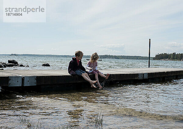 Bruder und Schwester sitzen auf einem Steg am Strand und paddeln mit den Zehen