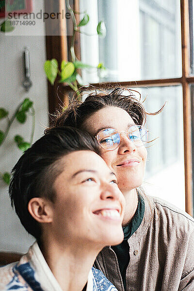 Süßes queeres weibliches Paar lächelt  während es zu Hause am Fenster kuschelt