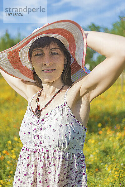 Porträt einer jungen schönen Frau mit Strohhut auf der Blumenwiese  während sie lächelnd in die Kamera blickt