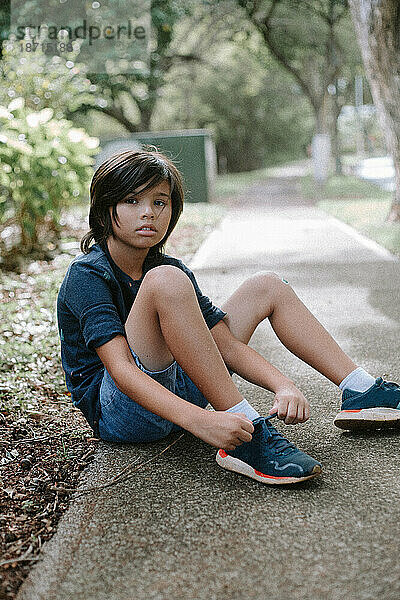 Junger  gemischtrassiger asiatischer Junge  der auf dem Bürgersteig sitzt und seine Schuhe bindet.