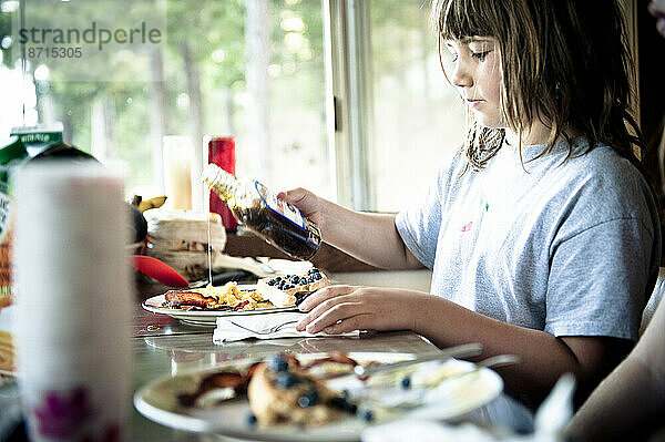 Ein junges Mädchen gießt Sirup über ihr Frühstück: Speck  Eier und Pfannkuchen.