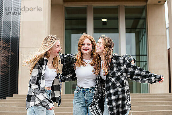 Drei lachende Teenager-Mädchen umarmen sich draußen auf Stufen.
