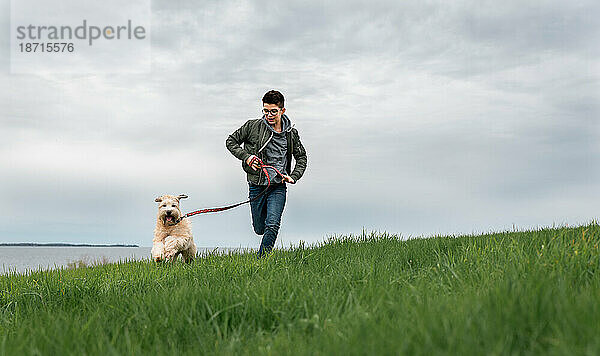 Teenager rennt an einem bewölkten Tag mit seinem Hund einen grasbewachsenen Hügel hinauf.