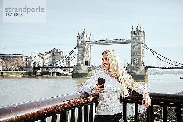 Attraktive Frau schaut entlang und hält ihr Mobiltelefon vor dem Hintergrund der Tower Bridge