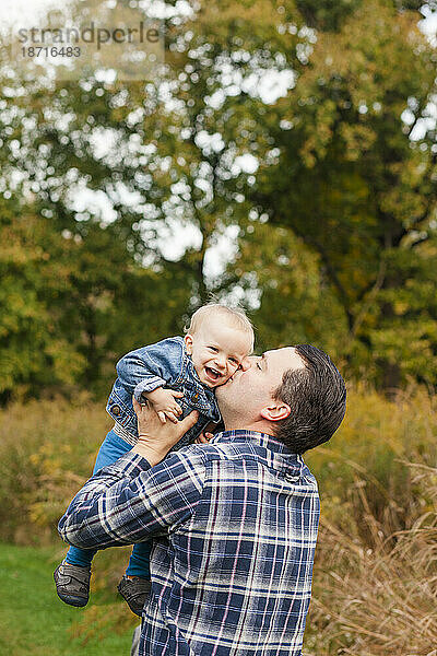 Ein glücklicher Vater streckt seinen lachenden kleinen Jungen in einem Park zum Kuss hoch