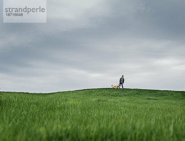Ein Junge und sein Hund stehen an einem bewölkten Tag weit entfernt auf einem Grashügel.