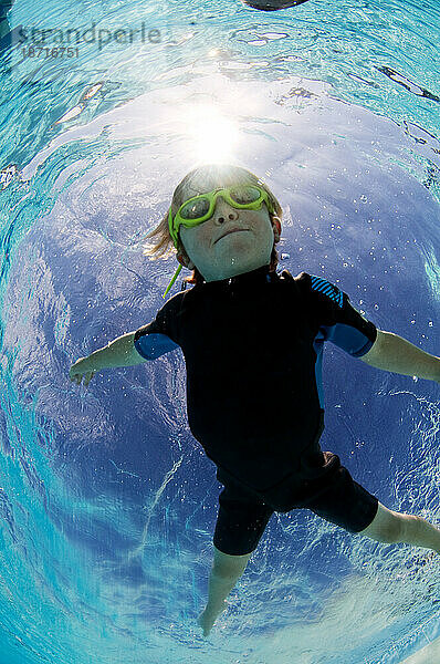 Unterwasseransicht eines kleinen Jungen  der eine Schutzbrille trägt und in einem Schwimmbad schwimmt.