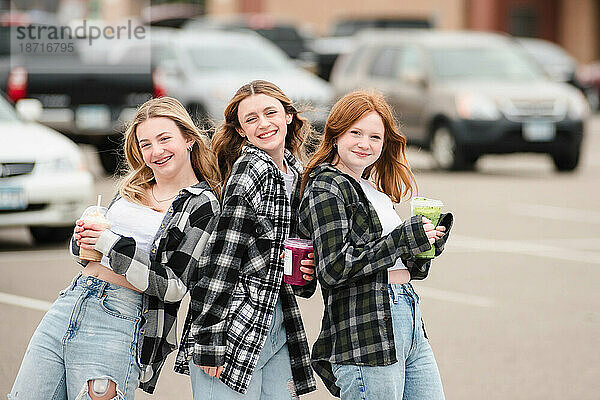 Drei glückliche Teenager-Mädchen halten Getränke auf einem Parkplatz.