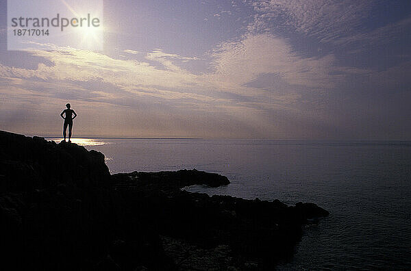 Eine Frau steht am Ufer des Cutler State Park  Cutler  Maine  während die Sonne über dem Meer aufgeht.